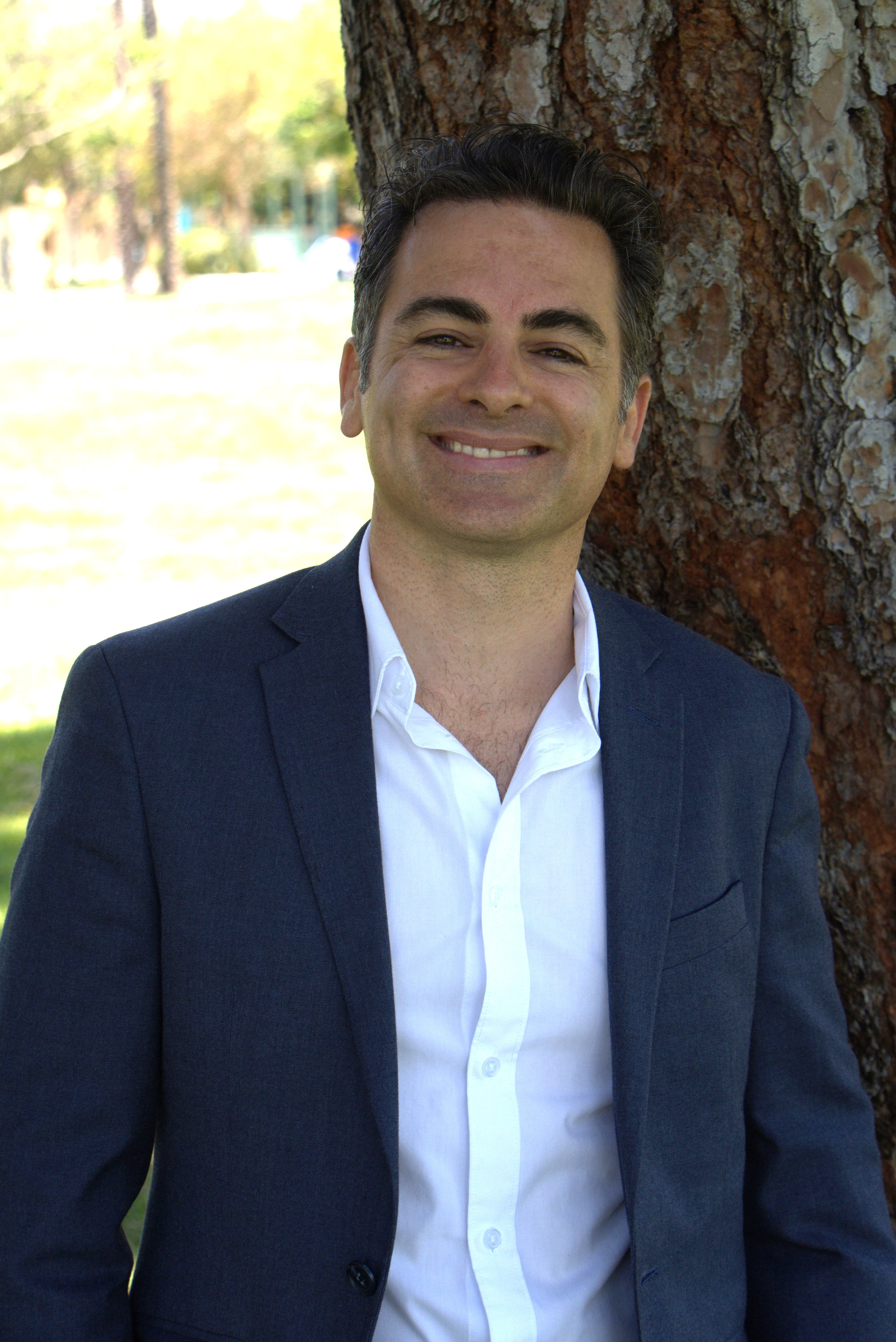 Smiling man in a navy blazer and white shirt leaning against a large tree outdoors.